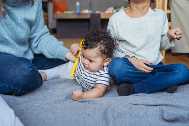 Baby sitting with a toy on a blanket beside an older brother and their mother — multi-stage baby toys that grow with your child