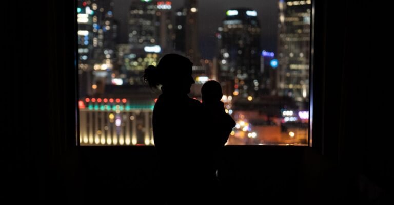 A mother holding her baby at night by a window overlooking a city skyline